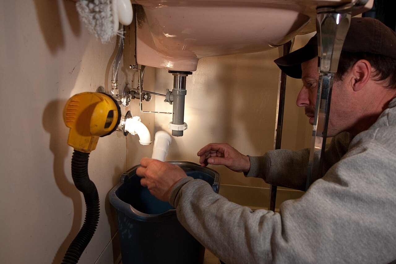 Un plombier intervient sous un lavabo, un seau posé sous le siphon